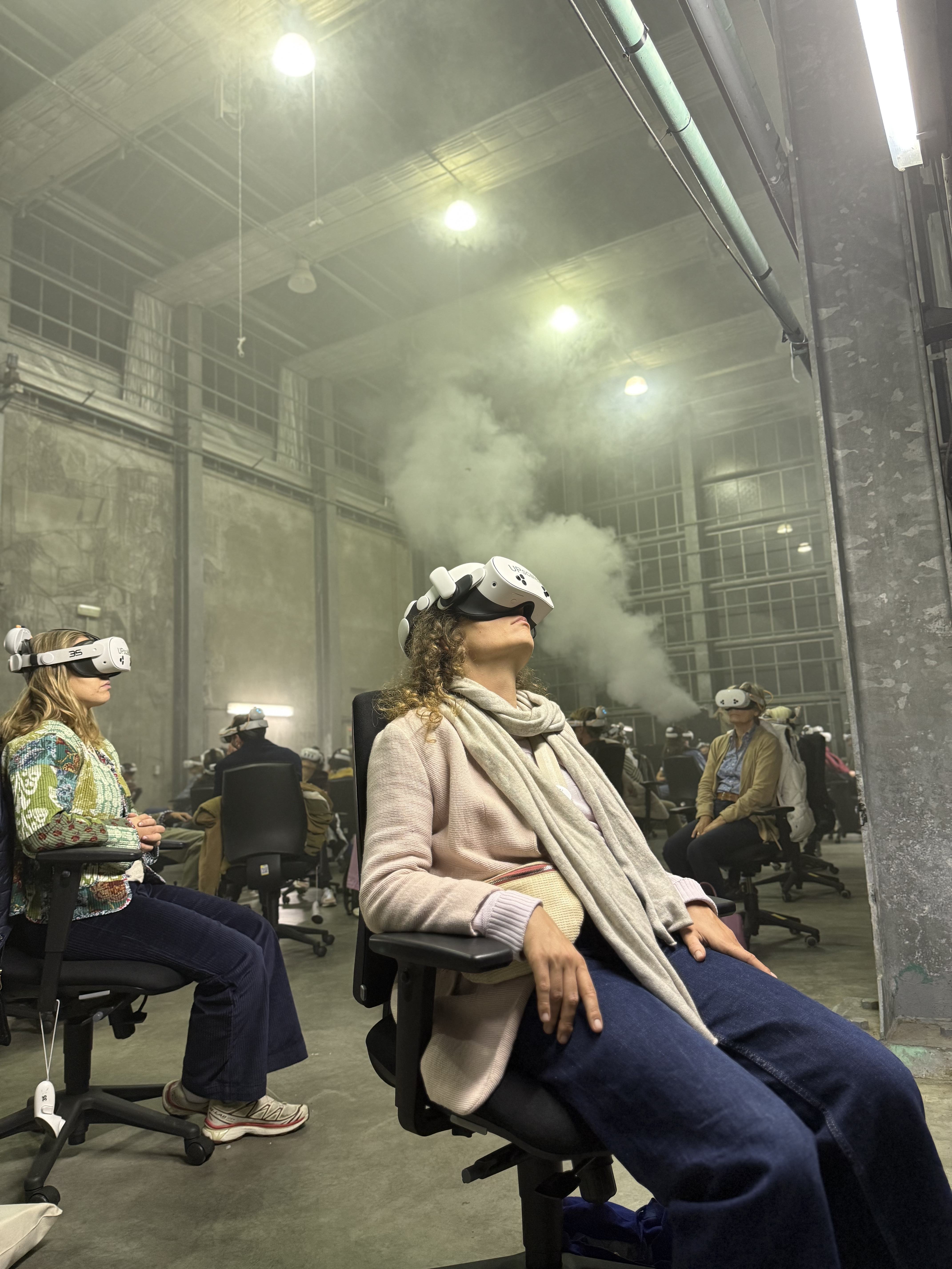 Verschillende mensen zitten op bureaustoelen door elkaar heen in een ruimte gevuld met rook, allen met een VR-bril op hun hoofd. Zittend persoon op voorgrond heeft hoofd iets naar boven getilt.