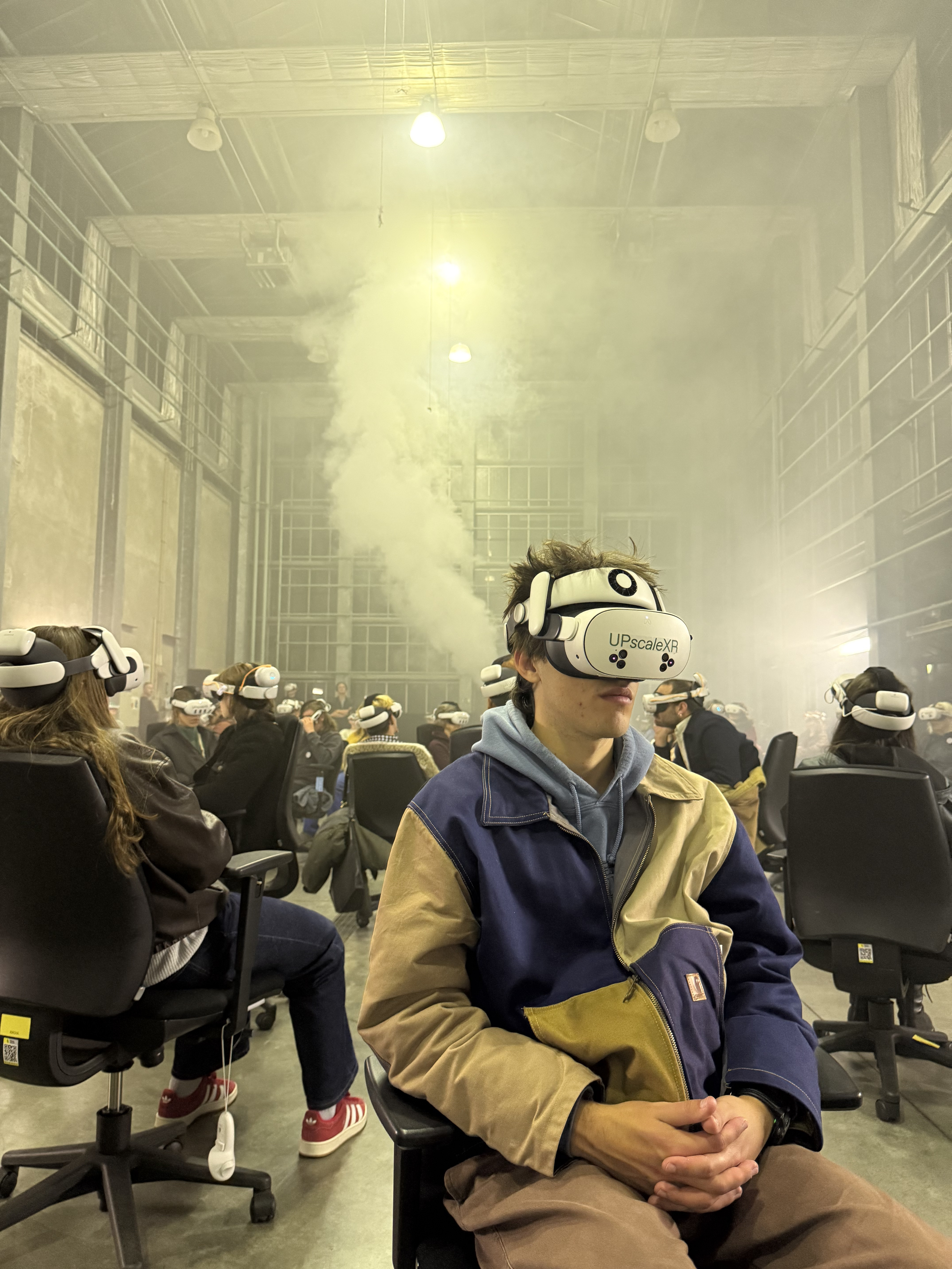 Verschillende mensen zitten op bureaustoelen door elkaar heen in een ruimte gevuld met rook, allen met een VR-bril op hun hoofd.