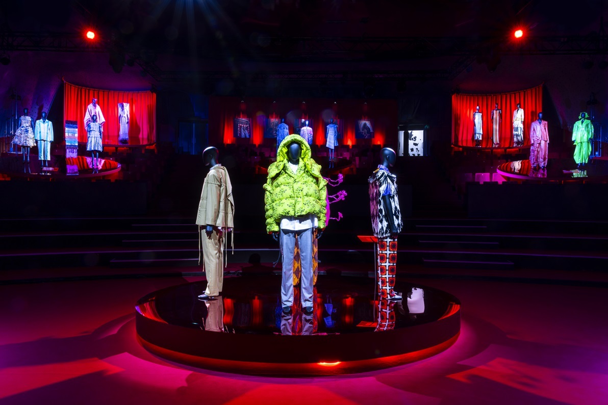 Rond podium in een ruimte met rode belichting op de vloer. Op het podium staan vier paspoppen met verschillende designkleren aan in verschillende kleuren.